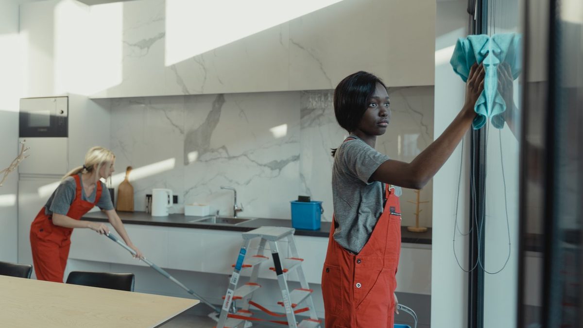 Cleaner in red overalls wiping a window in a bright modern apartment kitchen. Photo by Tima Miroshnichenko on Pexels