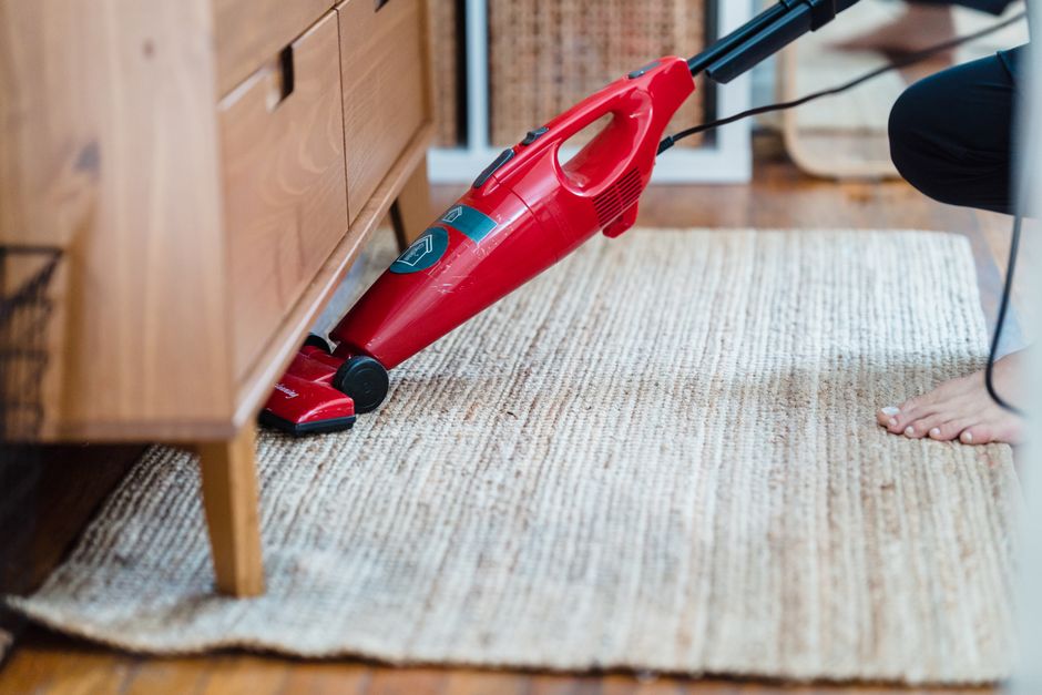 Side view of a person using a red vacuum under furniture on a rug for effective cleaning.