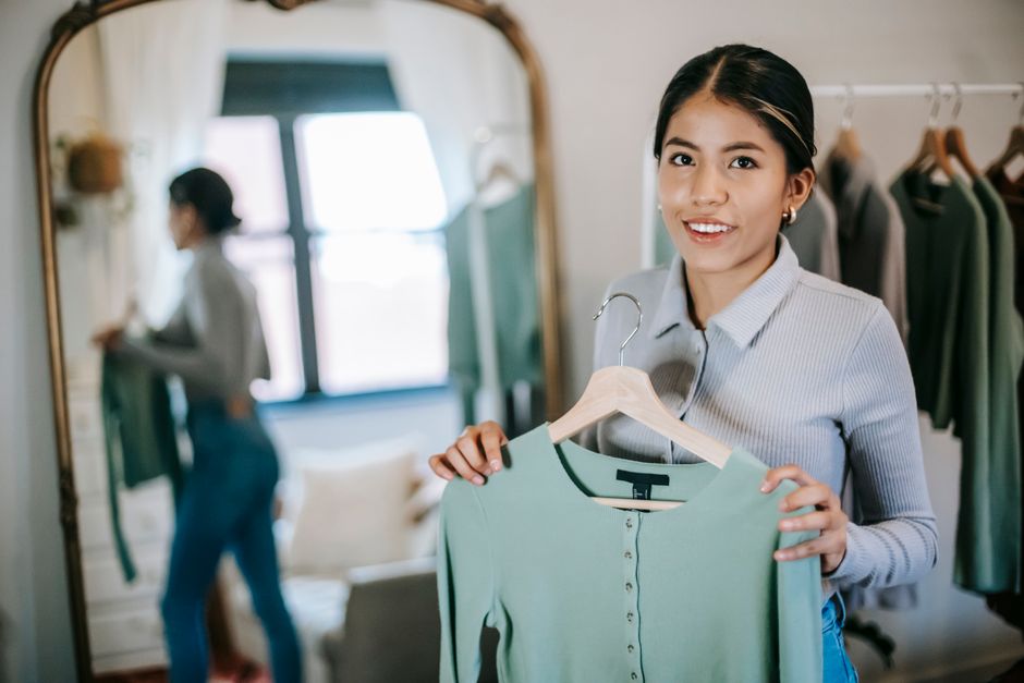Photo by Liza Summer on Pexels Smiling young ethnic woman in blouse and jeans standing with blue cloth on hanger at home near wardrobe and mirror while looking at camera