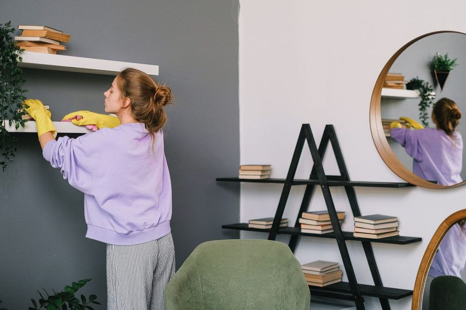 Woman in purple top cleaning living room shelves and a mirror in a bright apartment. Photo by SHVETS production on Pexels