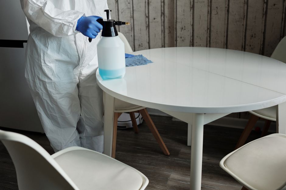 Person in protective suit disinfecting a white table indoors with a sprayer. Photo by Matilda Wormwood on Pexels