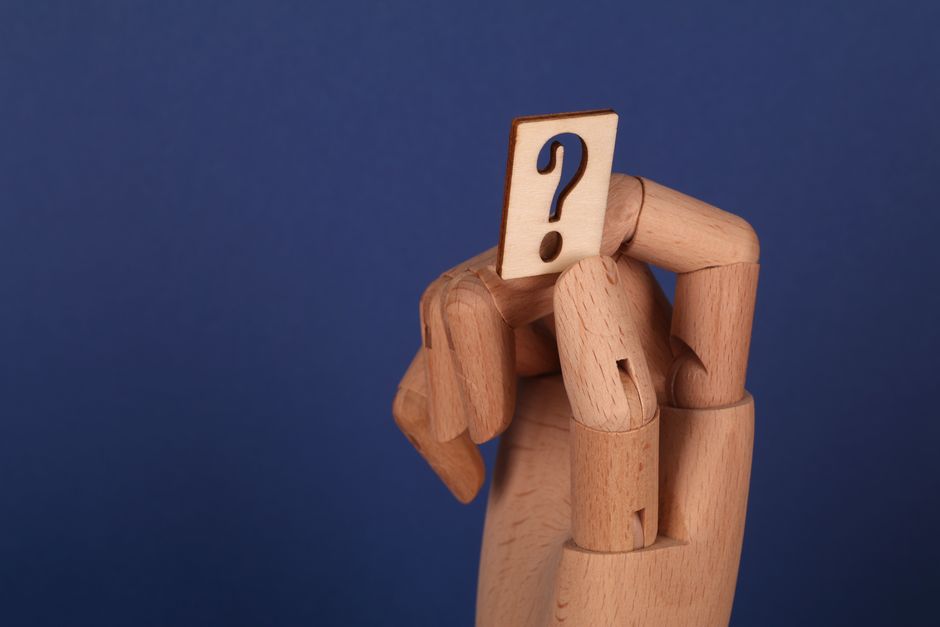 A wooden hand model holding a cut-out question mark against a blue background, symbolizing curiosity or inquiry. Photo by Ann H on Pexels