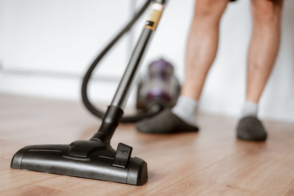 Person vacuuming a wooden floor with an upright vacuum cleaner in a bright room. Photo by Anete Lusina on Pexels