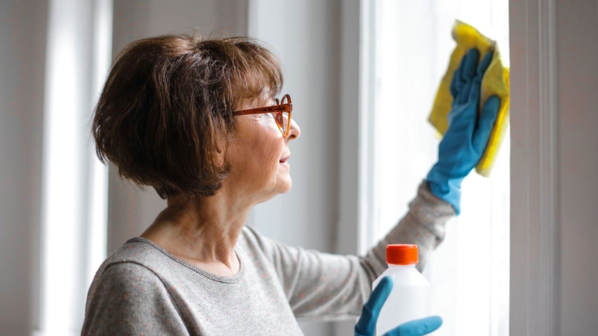 Woman wiping a window with a yellow cloth and spray bottle to remove dust in a condo. Photo by Andrea Piacquadio on Pexels