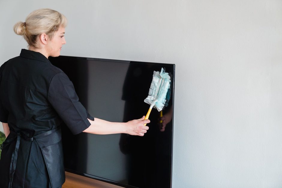 Woman in black dusting a wall-mounted panel with a duster in a white room. Photo by Liliana Drew on Pexels