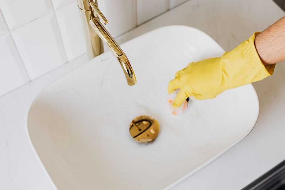 Yellow-gloved hand cleaning a white bathroom sink with brass tap. Photo by https://kaboompics.com/ on Pexels