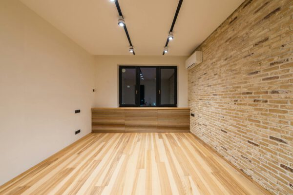 Empty modern room featuring wooden flooring, brick accent wall, and recessed lighting.