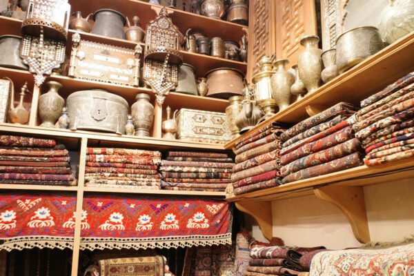 A vibrant scene showcasing folded Persian carpets and antique metalware in a traditional store setting.