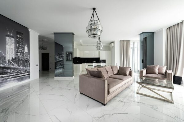 Interior of modern light studio with couch and armchair with pillows near glass table under chandelier next to open kitchen with white cabin