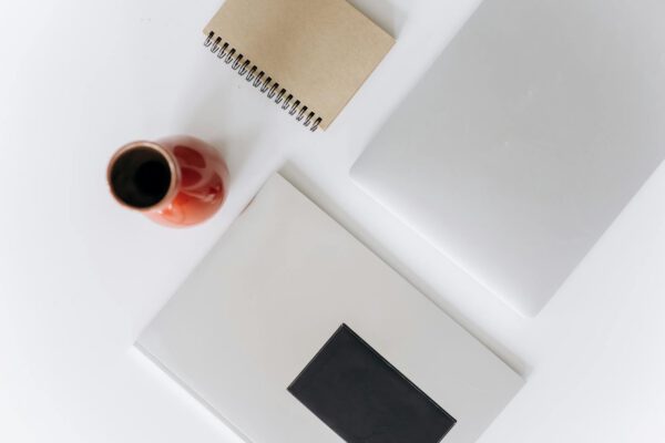 A top view of a minimalist office setup featuring a laptop, notebook, and red vase on a white table.