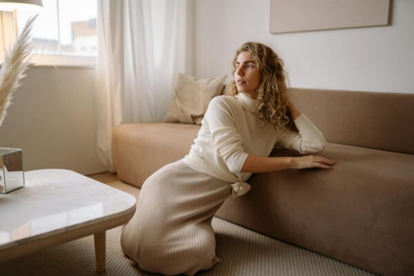 A woman in a cozy living room setting, relaxing beside a beige sofa with natural light.