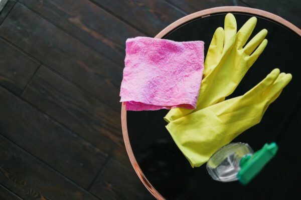 From above of yellow latex gloves placed near rag and spray detergent on table against wooden floor in daytime