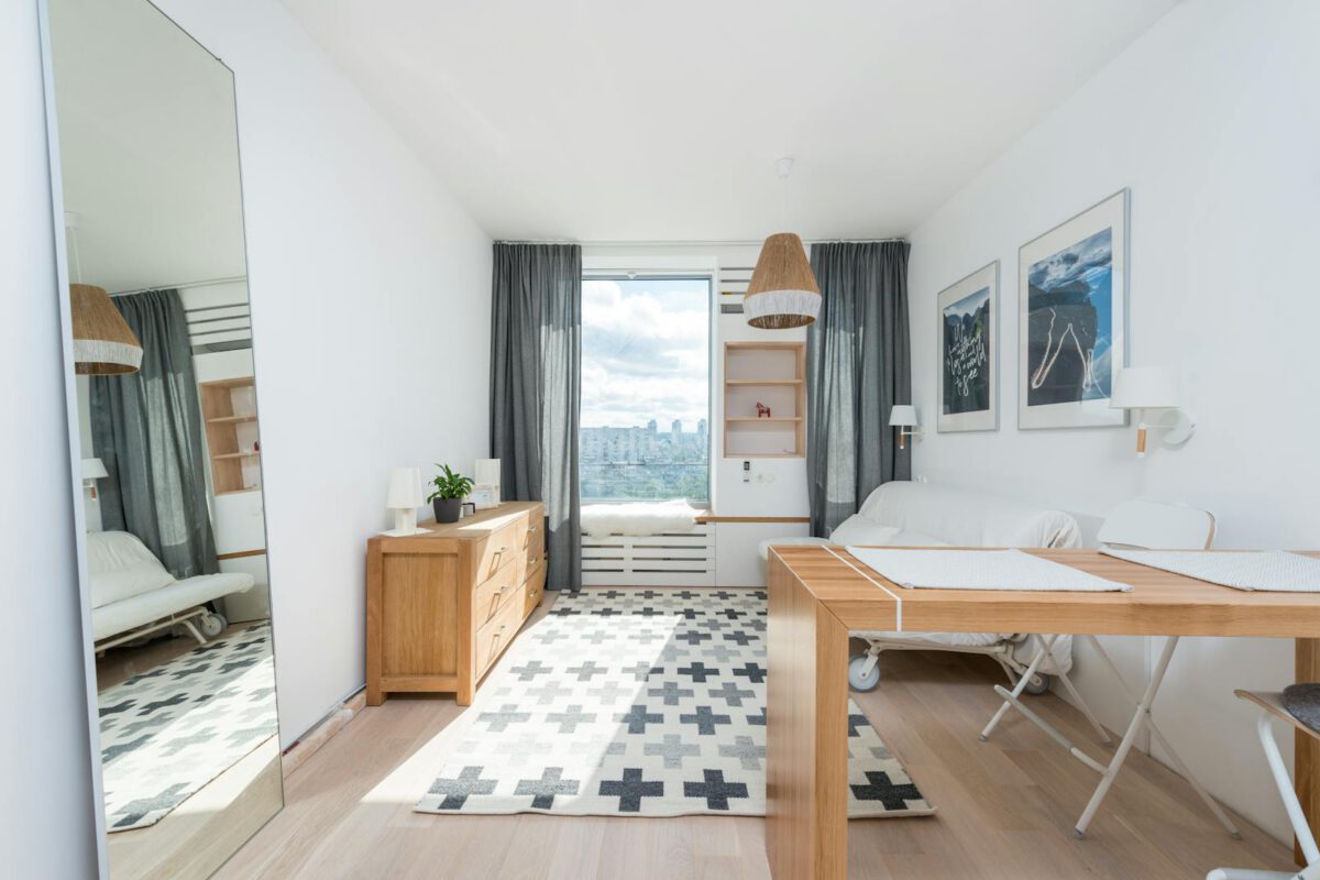 Wooden table and drawer placed in room with big window and wall decorated with pictures in light room with mirror