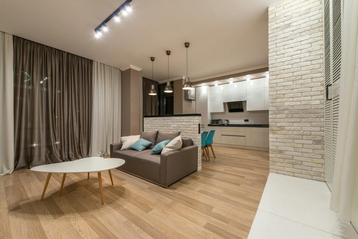 Comfortable sofa with cushions near table in stylish living room with curtains on window near kitchen zone with modern appliances
