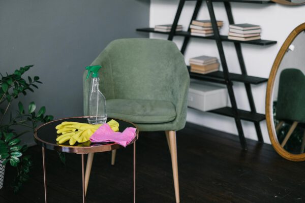 Yellow latex gloves placed near spray detergent bottle and rag on round table against comfortable chair in living room in daytime