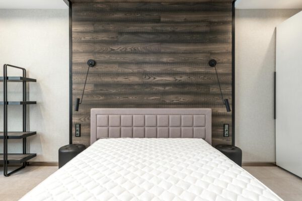 Comfortable bed with white mattress placed at wall with wooden element in stylish spacious bedroom with lamps and shelves in apartment