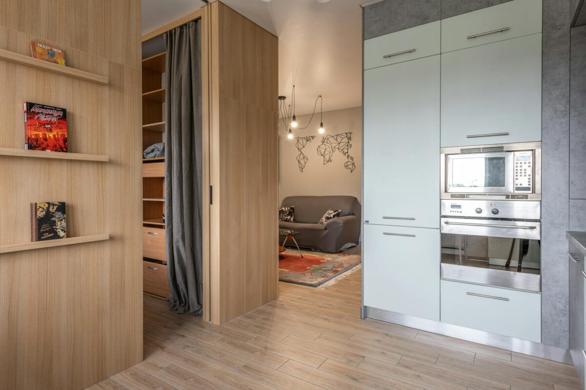 Modern apartment with cabinets with oven and microwave in kitchen near wooden shelves with books near entrance in room with wardrobe with cu