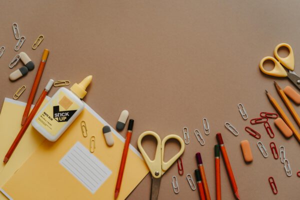 Flat lay of assorted stationery items like scissors, pencils, and paper clips on a brown background.