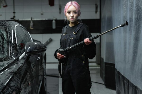 Asian woman with pink hair using a pressure washer at an indoor car wash.