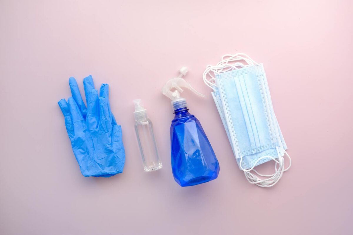 Flat lay of protective gear including gloves, sanitizer, and masks on a pastel pink background.