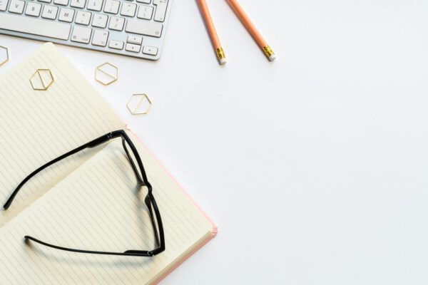 A clean and minimalist home office setup with glasses, notebook, keyboard, and pencils on a white background.