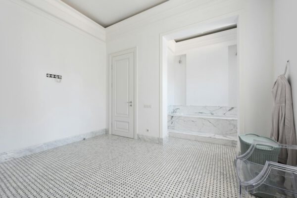 Spacious white bathroom with marble decor, featuring a bathrobe and modern chair.