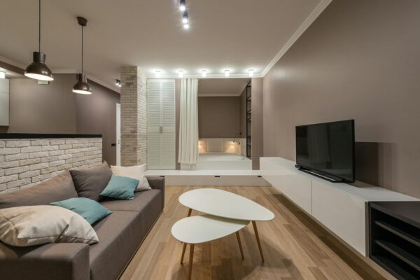 Comfortable gray sofa with colorful cushions placed near table and modern TV on white cabinet in spacious living room with tub