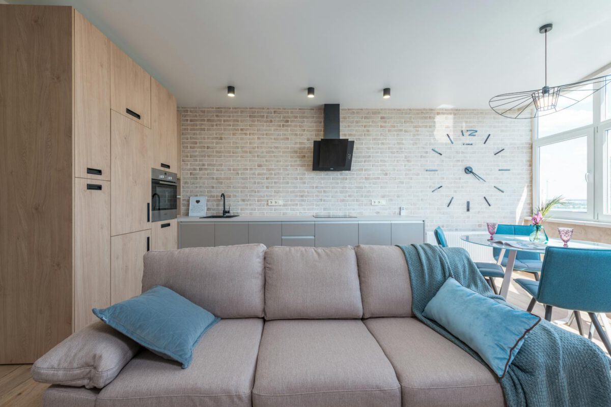 Minimalistic interior design of modern light studio consisting of big sofa and kitchen with dining table and chairs against brick wall in da