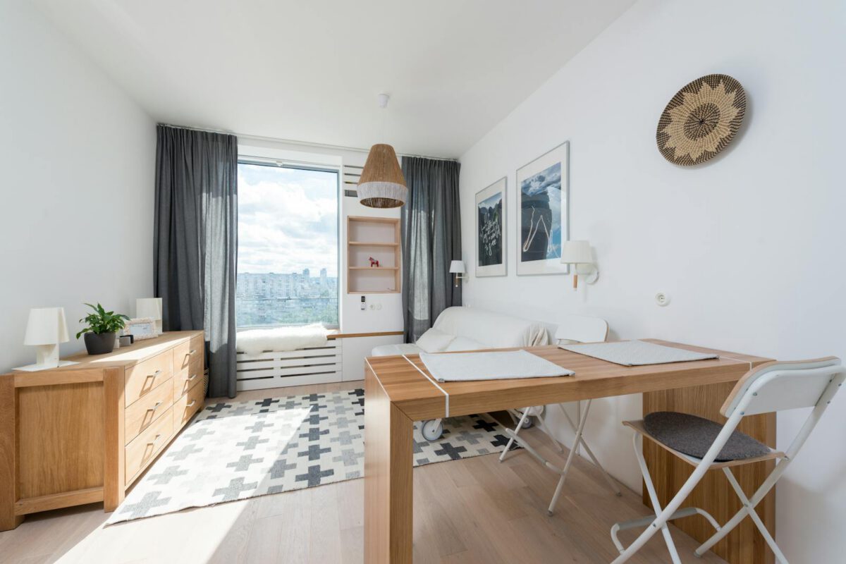 Table with chair placed against window with curtains in stylish room with wooden drawer and wall decorated with creative pictures