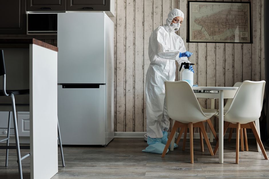 Person in hazmat suit disinfecting a modern kitchen table with spray bottle for hygiene.
