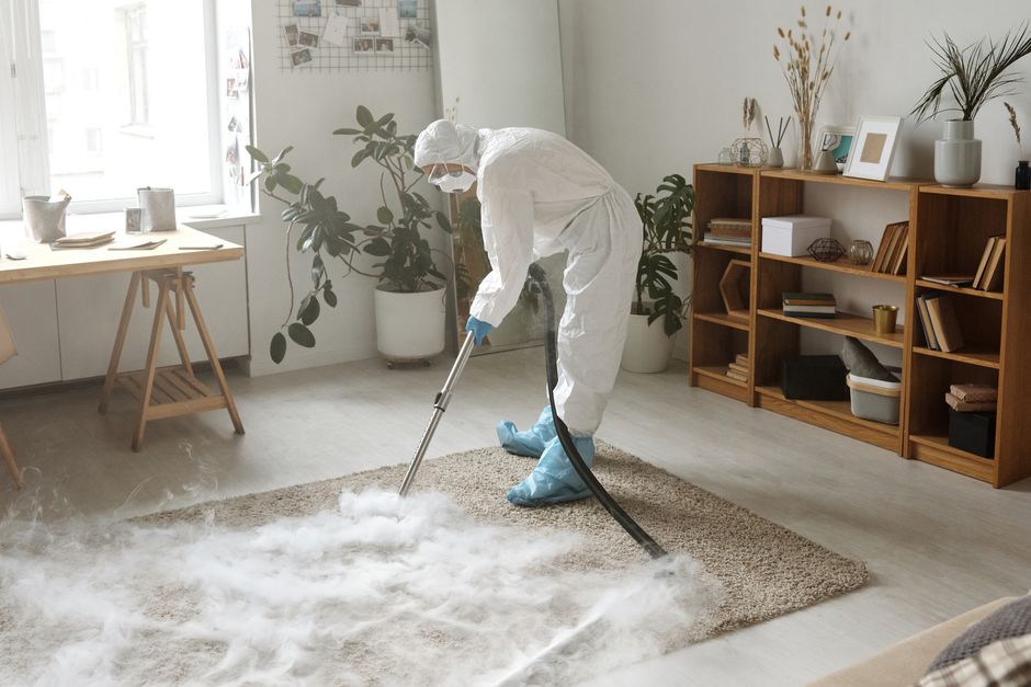 A woman in protective gear disinfects a carpet indoors to ensure safety during the COVID-19 pandemic.