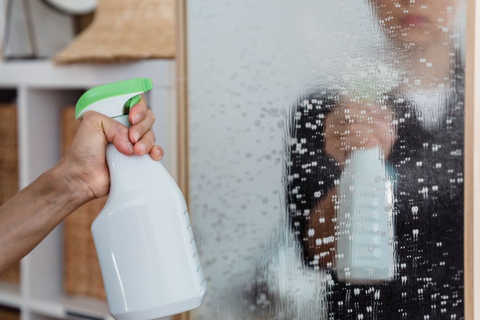 Close-up of a hand using a spray bottle to clean a mirror indoors.