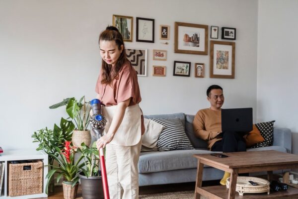 An Asian woman vacuuming while a man works on a laptop in a modern living room.